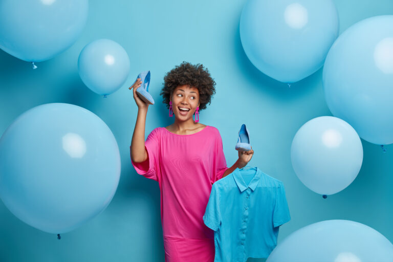Photo of beautiful cheerful woman chooses what to wear, selects blue outfit for special event, holds high heel shoes and shirt on hanger, poses against decorated background. Clothes collection